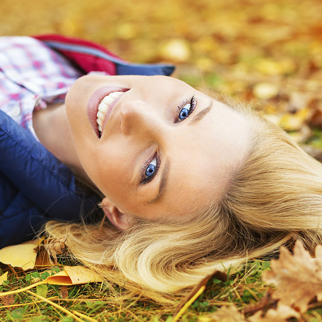 Blonde Frau liegt auf einer Wiese mit Herbstlaub. Man sieht nur den Kopf, von der Stirn aus.